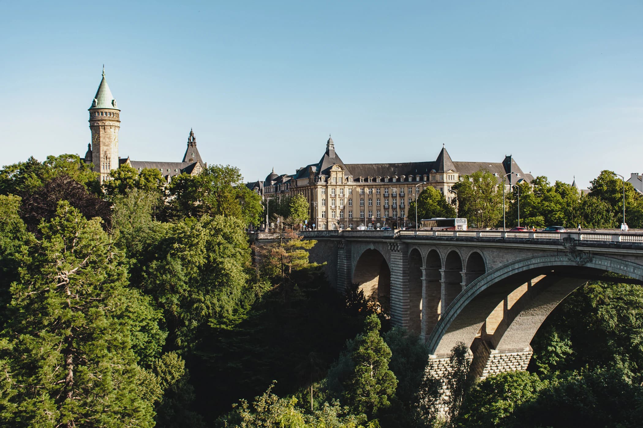 The historic Adolphe Bridge spanning a lush green valley in Luxembourg City, with the State Savings Bank tower in the background.