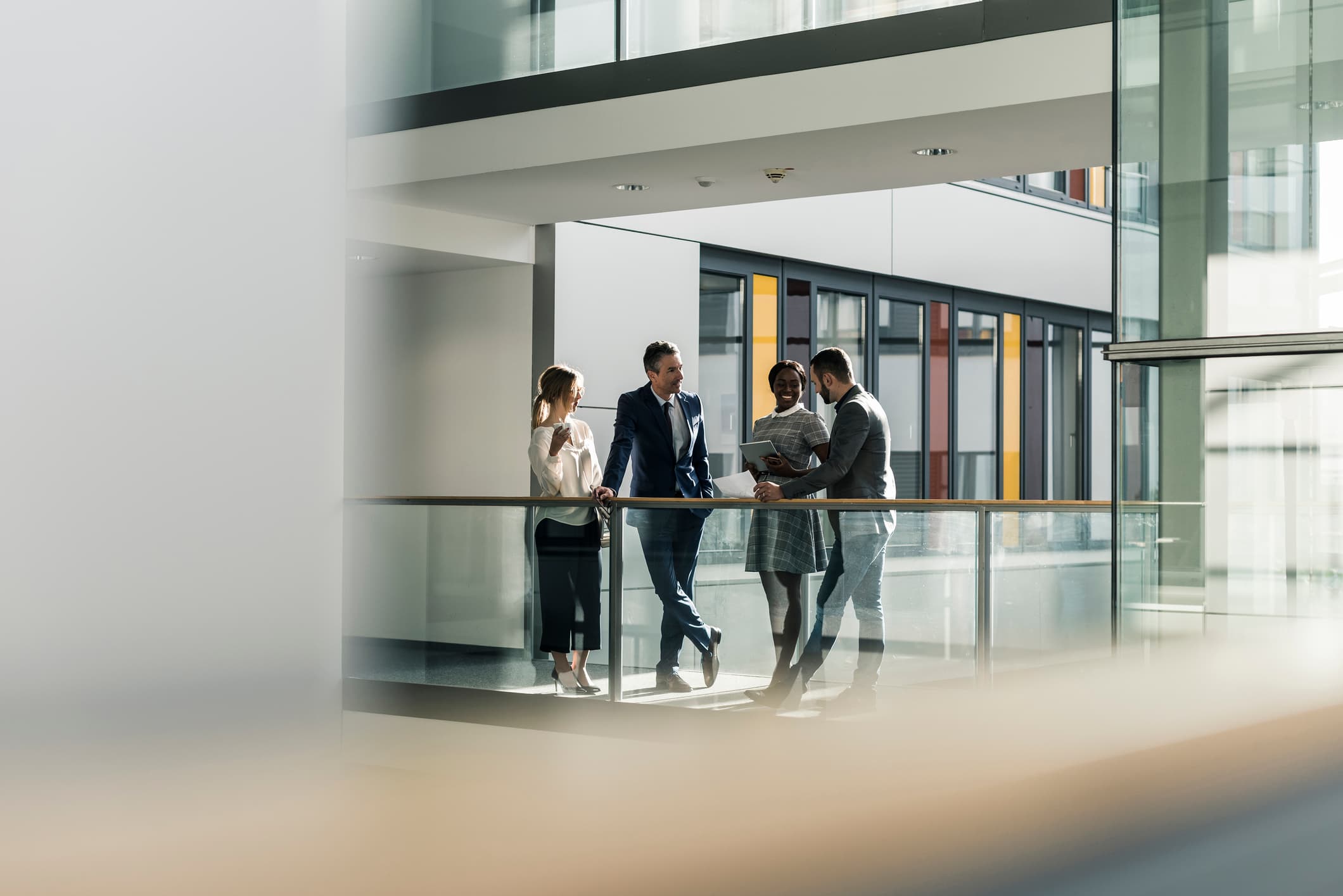 Four diverse coworkers talking on an indoor balcony walkway of a modern, multi-story office building.