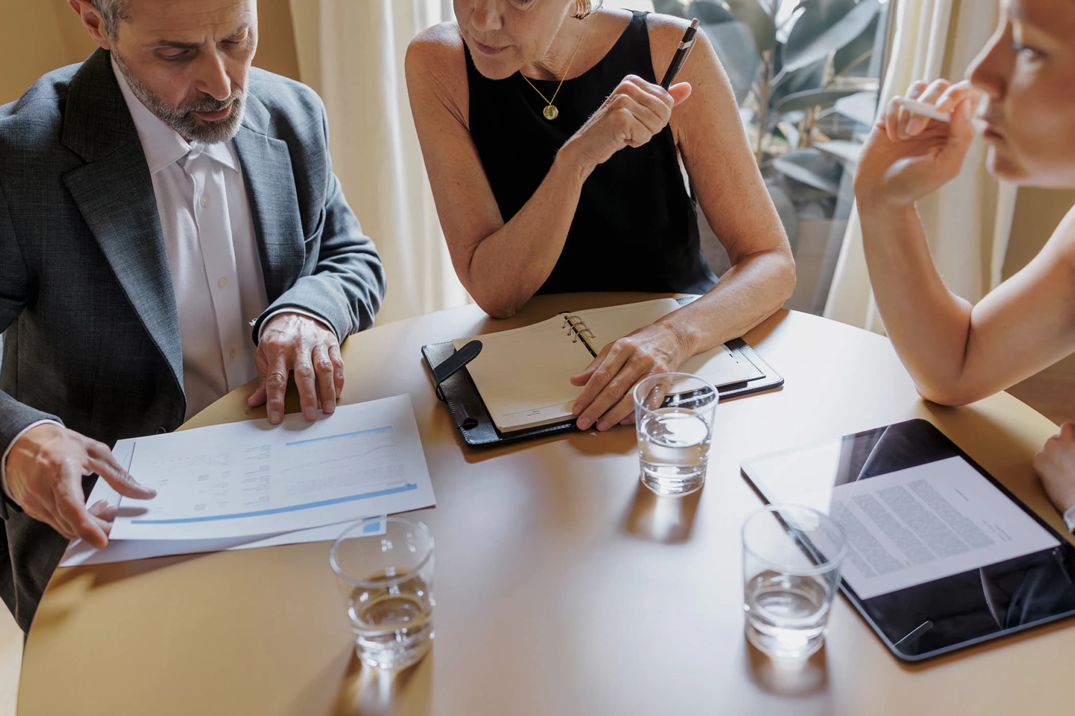 A close-up of three business professionals sitting at a round table, reviewing a printed document and a digital tablet.